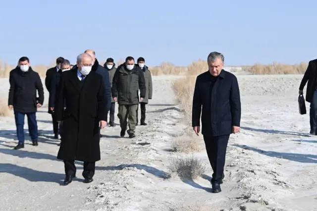 Prezident Orolning qurigan tubida, cho‘llangan va sho‘rlangan yerdan yurib kelyapti, uning atrofida rasmiylar, maska taqqan. Hamma qishki kiyimda. Oftob charaqlab turgan kun.
