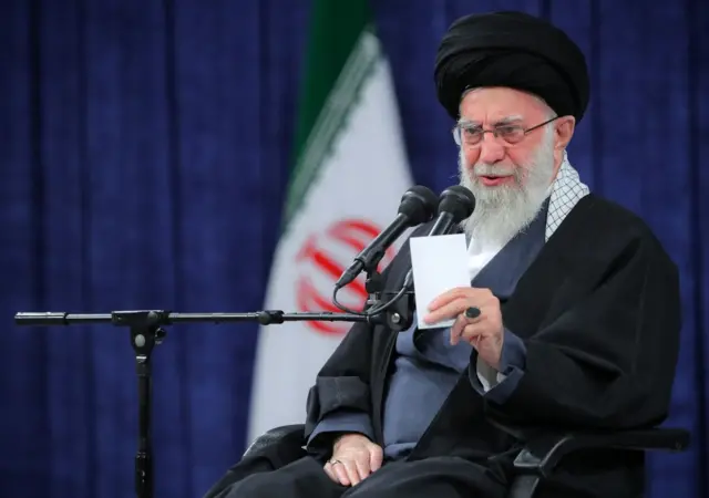 El ayatolá Alí Jamenei, vestido con ropa negra, está sentado sosteniendo un papel blanco en la mano frente a unos micrófonos, atrás se ve la bandera de Irán