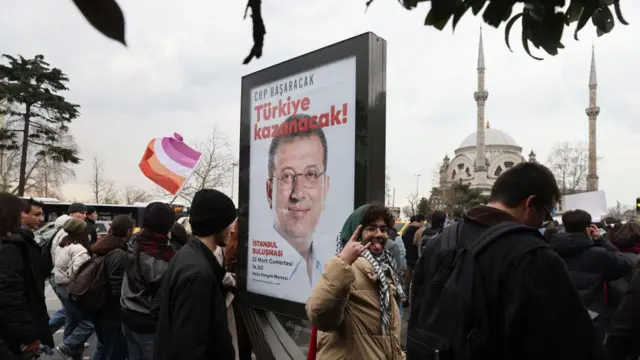 İmamoğlu posterinin önünde bir genç, kameraya dil çıkarıyor. ARkdasında kalabalık ve bir cami