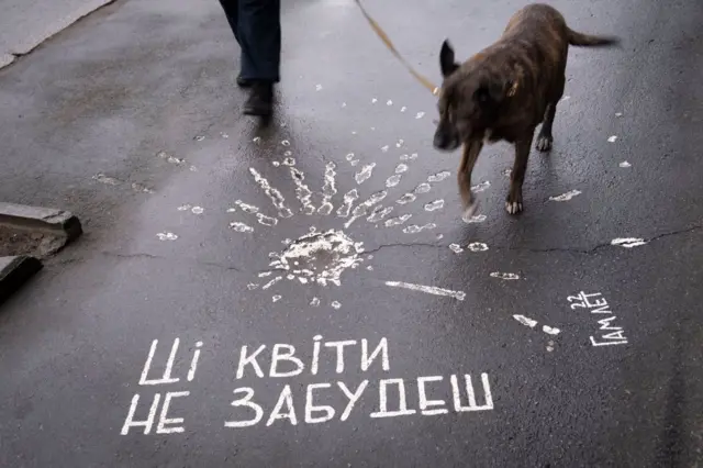 На фото зображено мокрий асфальт із білим малюнком у вигляді квітки, створеної з відбитків пальців, та написом : "Ці квіти не забудеш". Праворуч підпис: "Гамлет". По асфальту йде людина, поруч із нею – собака на повідку.