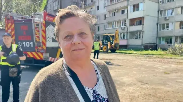 茶色のカーディガンと花柄のシャツを着たショートヘアのヴァレンティーナさんが、アパートの前に立っている。背後には消防士と消防車が見える