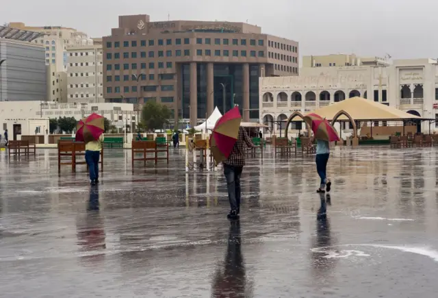 يتجول 3 أشخاص، بمظلات في سوق واقف، خلال يوم ماطر في الدوحة- قطر. تظهر مجموعة من المباني، والأمطار تشكل تجمعاً من المياه.