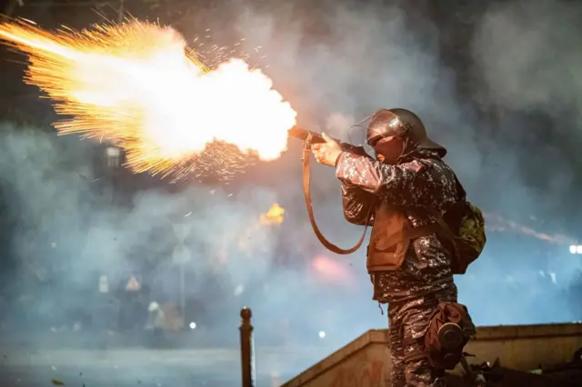 Agente de la policía vestido con camuflaje militar, casco y chaleco antibalas con un arma larga en la mano de la que sale una enorme llamarada tras apretar el gatillo y dejar salir una bomba lacrimógena. 