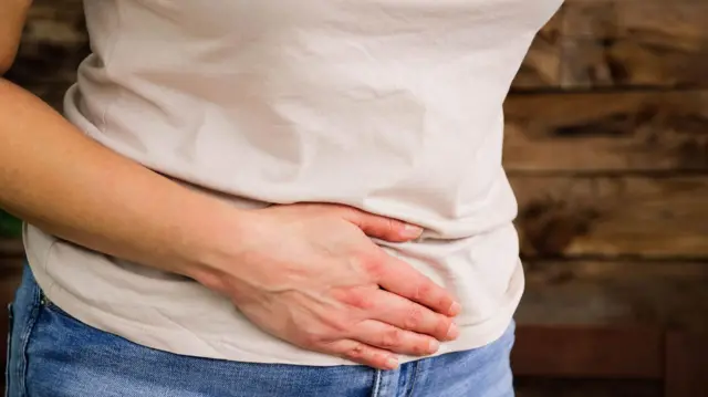 Fotografie de stoc: o imagine în prim plan a unei femei cu mâna pe stomac. Ea poartă un tricou roz deschis și blugi albaștri.