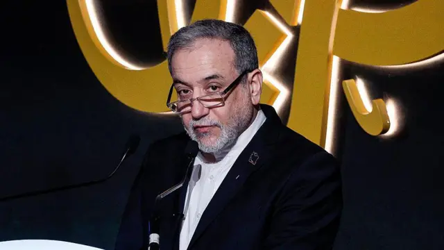 Koyu mavi bir ceket, beyaz gömlek, gözlük ve kol saati takan Abbas Araghchi, 7 Şubat 2026'da Doha'da düzenlenen 17. El Cezire Forumu'nda bir kürsüden mikrofona konuşuyor.