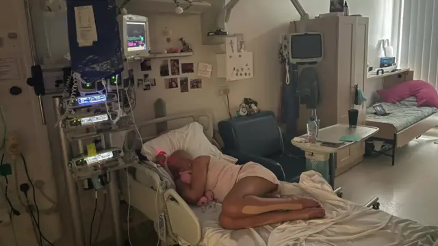 La Sra. Cassell, quien perdió el cabello durante el tratamiento, está acostada de lado en una cama de hospital, abrazando a un osito de peluche rosa. Está rodeada de equipo médico.