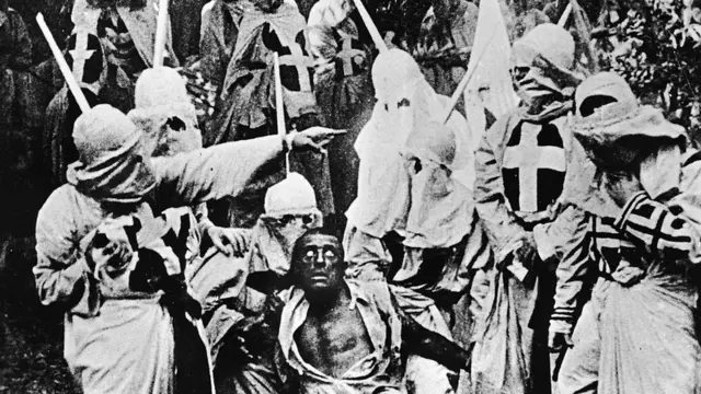 Ку-клукс-клан в полном парадном облачении готовится линчевать чернокожего. Кадр из фильма «Рождение нации». Роль чернокожего играет белый актер с выкрашенным черной краской лицом.