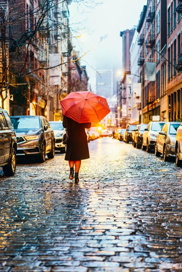 من الخلف شابة تمشي تحت المطر، ترتدي ملابس شتوية تبدو سوداء اللون، وتحمل مظلة حمراء في نيويورك.