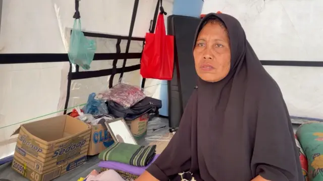 Armiayati menggunakan kerudung panjang berwarna hitam, duduk di dalam tenda pengungsian berwarna putih. Di belakangnya ada barang-barang seperti kardus, kantong plastik yang digantung, tumpukan baju, dan beberapa bantal.