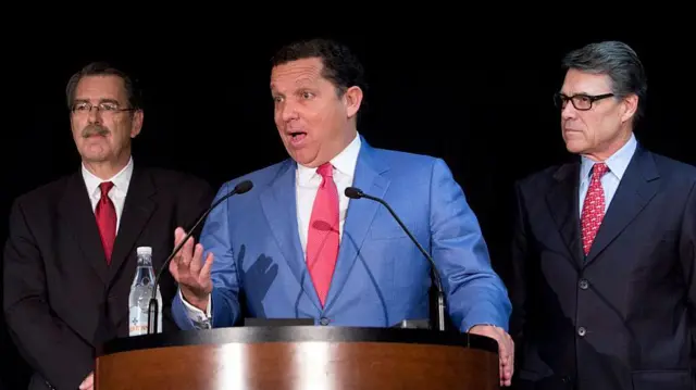 Di lawyer Tony Buzbee, wear a light blue suit, stands at a podium during a press conference, as former Texas govnor Rick Perry watch from im left hand side