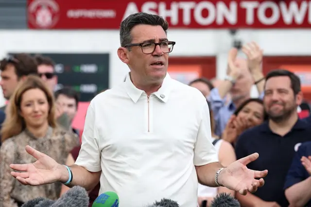 Burnham, vêtu d'un polo blanc, ouvre les bras, avec une foule derrière lui