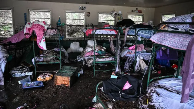 泥まみれになった2段ベッドが並ぶ。ベッドにはピンクやパープル、花柄などのシーツや掛布団が見える。泥まみれのバッグや枕なども散乱している
