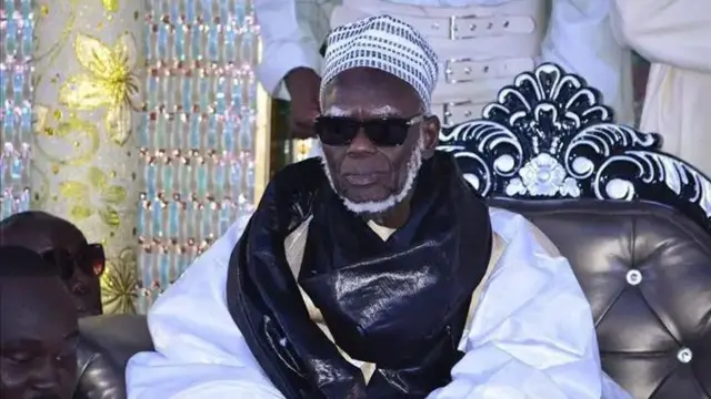  Serigne Mountakha Bassirou Mbacké zaune kan kujera sanye da fararen tufafi da baƙin mayafi anye da baƙin gilas da hula