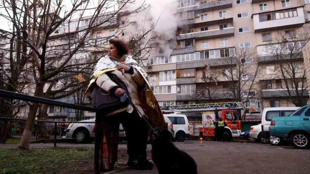 Yardım battaniyesini sarılmış bir kadın, bir eliyle köpeğinin tasmasını tutuyori sol yana bakıyor. Arkada, dumanlar tüten bir binanın önünde itfaiye aracı var. 