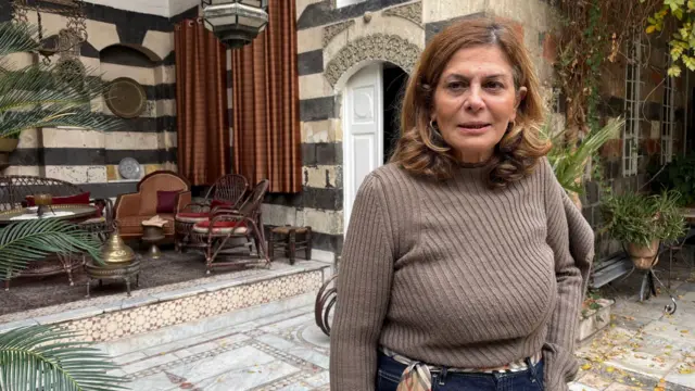 قبوات في حديقة منزلها، وهو منزل سوري تقليدي مبني من الطوب الفاتح والداكن، بأبواب خشبية بيضاء، وبلاط مزخرف على الدرج، ونباتات متسلقة حول النوافذ. ترتدي قبوات كنزة بنية فاتحة اللون، وتنظر قليلاً إلى يسار الكاميرا بنظرة متأملة.