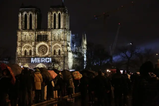2024 年 12 月 7 日,法國巴黎,在巴黎聖母院重新開放儀式上,法語單字「MERCI」投射在巴黎聖母院正面。門外有一批人在排隊。