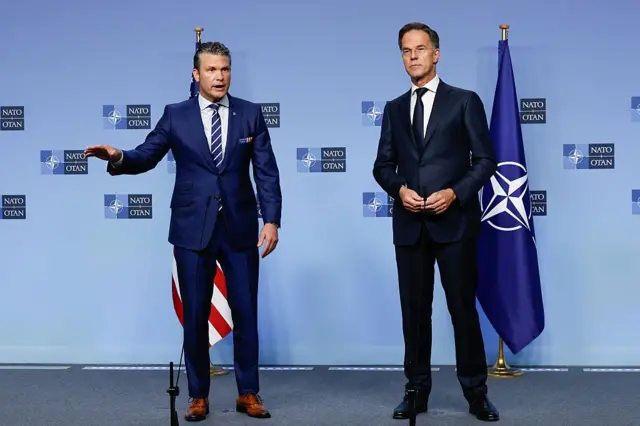 ABD Savunma Bakanı ve NATO Genel Sekreteri mavi bir fonda yan yana basına konuşuyorlar. İkisi de ayakta