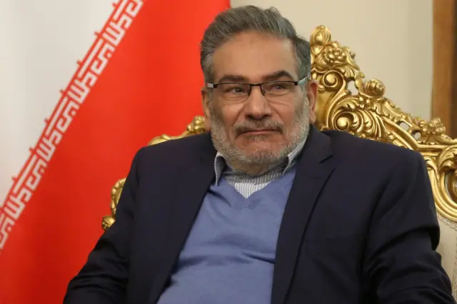 Com jaqueta azul escura, colete azul claro, camisa listrada e óculos, o contra-almirante Ali Shamkhani está sentado em uma cadeira dourada em frente a uma bandeira iraniana, durante uma reunião com o conselheiro de segurança nacional dos Emirados Árabes Unidos, Tahnoun bin Zayed Al Nahyan, em Teerã, no dia 6 de dezembro de 2021