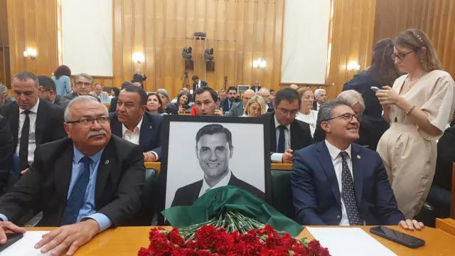 Ferdi Zeyrek'in siyah beyaz çerçeveli resmi önünde karanfillerle Meclis sıralarında milletvekillerinin arasında oturuyor. 