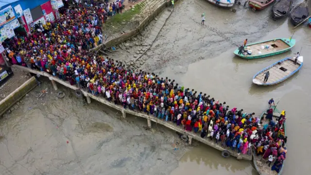 बांग्लादेश के मोंगला शहर में पोशुर नदी को नाव से पार करते हुए जलवायु प्रवासी मज़दूरों की भीड़ का हवाई दृश्य, जो समय पर अपने कार्यालय पहुँचने की जल्दी में हैं. कई प्रवासी मोंगला के बंदरगाह शहर में बेहतर जीवन बनाने और मज़बूत बनने के लिए निर्यात प्रसंस्करण क्षेत्र (EPZ) में काम करते हैं.

