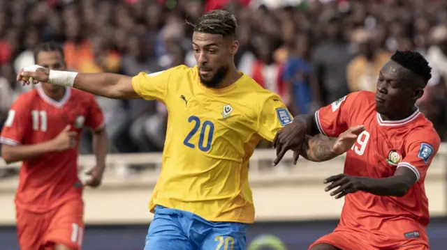 Denis Bouanga amevalia shati la manjano la Gabon lenye namba 20 kifuani na kaptura ya bluu huku akinyoosha mikono yake kusawazisha na kulinda mpira wakati wa mechi ya soka kutoka kwa beki wa Kenya Rooney Onyango, ambaye amevalia shati jekundu. Mchezaji wa pili wa Kenya anaweza kuonekana nyuma akikimbia kuelekea wawili hao huku nyuma yake unaweza kuona stendi iliyojaa wafuasi.