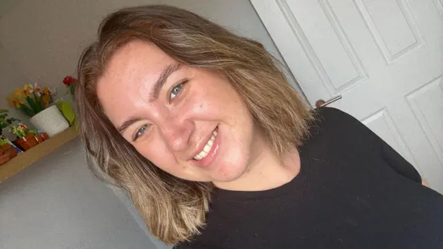 Gabby gülümseyerek selfie çekiyor. Siyah bir tişört giymiş ve omuz hizasında kahverengi saçları var.