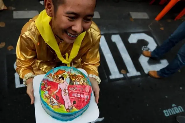 น.ส.จตุพร แซ่อึง สมาชิกกลุ่มวีโว่ที่ถูกตั้งข้อหา 112 กับเค้กที่ผู้สนับสนุนนำมามอบเป็นกำลังใจ โดยตกแต่งหน้าเค้กเป็นรูปที่เธอสวมชุดไทยสีชมพูเดินพรมแดงในงานศิลปะคณะราษฎรเมื่อวันที่ 29 ต.ค. 2563