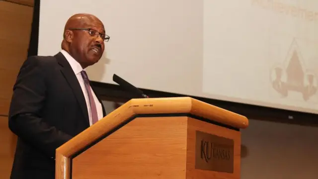Ngondi Kamatuka hablando en un podio durante una conferencia en la Universidad de Kansas donde trabaja