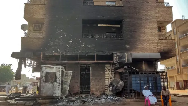 Bâtiment carbonisé à la suite d'une attaque, le rez-de-chaussée étant complètement détruit. Deux hommes sont assis sur des chaises colorées au pied du bâtiment.