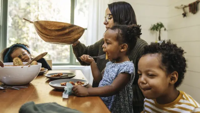 Mulher negrabônus de registro grátiscabelo preto e óculos segura uma tigelabônus de registro grátisfrente a uma mesa enquanto três crianças negras comem