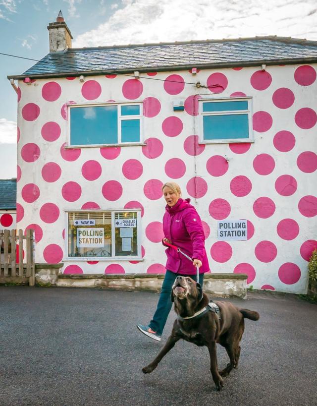 A woman and her dog walk past a polling station building that is covered in pink polka dots