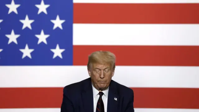 Trump olha para baixo, com bandeira dos EUA ao fundo