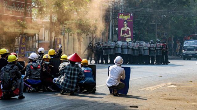 Protesters sit in a street holding makeshift shields as they face police in riot gear, in Mandalay on 3 March 2021