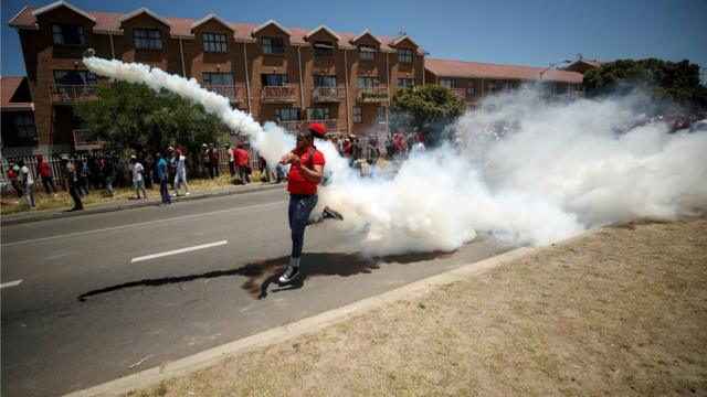 الغاز المسيل للدموع يملأ الجو، بينما يقوم متظاهر ضد تقارير تتحدث عن ممارسة العنصرية في إحدى مدارس كيب تاون في جنوب أفريقيا بإعادة رمي قنبلة غاز على الشرطة التي أطلقتها.
