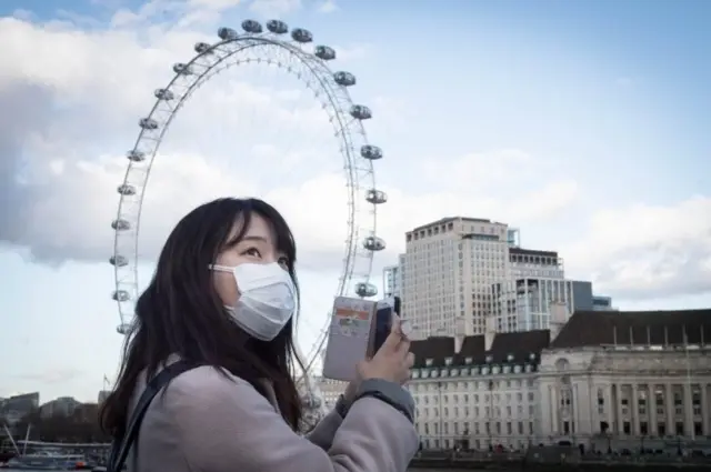 A woman wearing a face mask takes a photo on her phone from Westminster Bridge in London