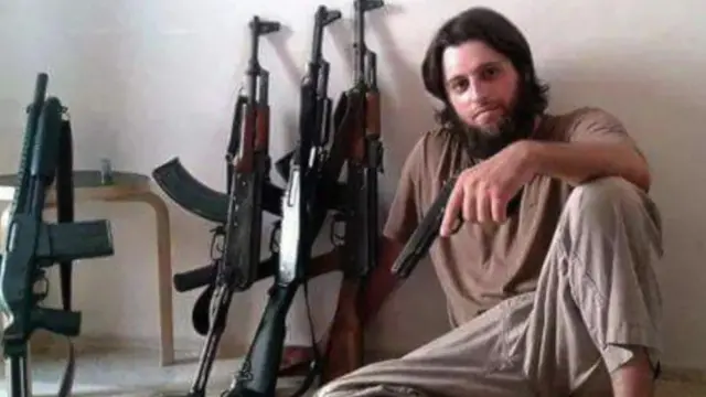 John Georgelas sits on a floor with assault rifles leaning on a wall next to him