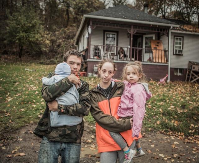 Greg y Ellen holding con sus dos hijos frente a una casa