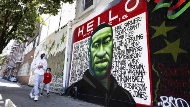 Pai e filha, negros e de máscara, passam em frente a grafite com retrato de Floyd e nome de outras pessoas vítimas da violência policial e do racismo