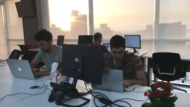 Two young men working at computers with the sunsetting out of the window of the skyscraper