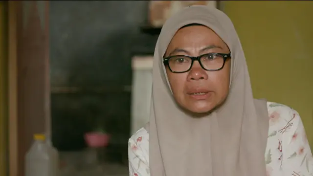 Rusehan mengenakan jilbab cokelat dan baju putih motif bunga. Dia mengenakan kacamata dengan bingkai warna hitam.