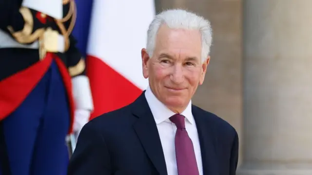 Ambasador Stanów Zjednoczonych we Francji, Charles Kushner, na zdjęciu uśmiecha się przed Pałacem Elizejskim w Paryżu. Ma białe włosy i ubrany jest w ciemny garnitur, białą koszulę i śliwkowy krawat.