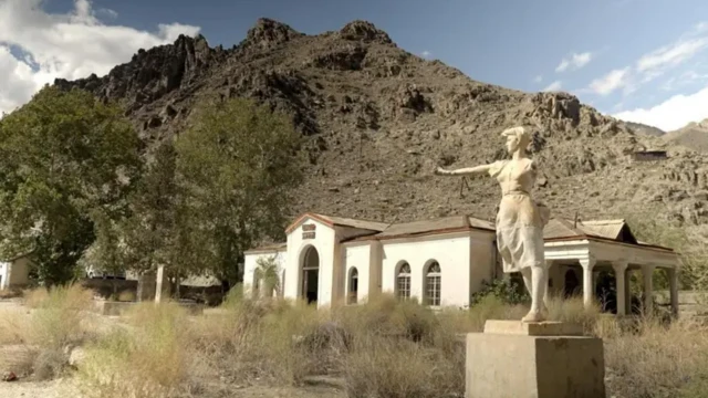 یک عمارت سفید رنگ متروکه در کنار کوه و یک مجسمه زنی که دستش شکسته است اطراف بوتههای خشک روییده است