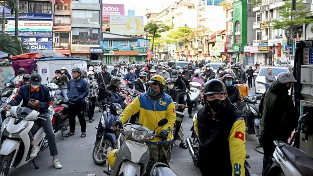 A crowd of people on motorcycles waiting on the road outside a petrol station, with the photo taken from inside the petrol station.