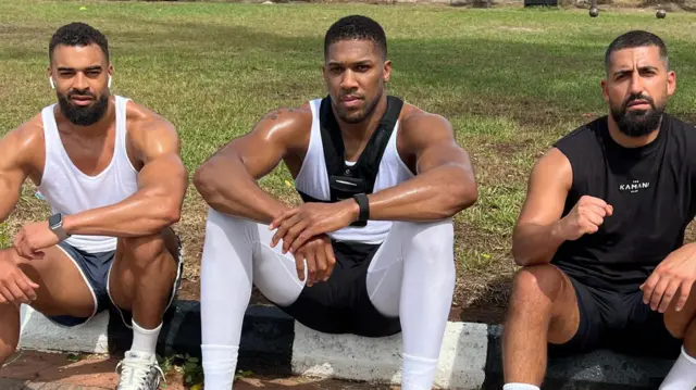 Latif Ayodele se sienta con Anthony Joshua y Sina Ghami en un bordillo con césped detrás, todos con ropa deportiva. Latif lleva auriculares blancos, una camiseta blanca y pantalones cortos azules. Tiene los brazos muy musculosos y lleva bigote y barba.