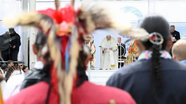 Francisco discursa ao fundo observado por pessoas com adereços indígenas coloridos.