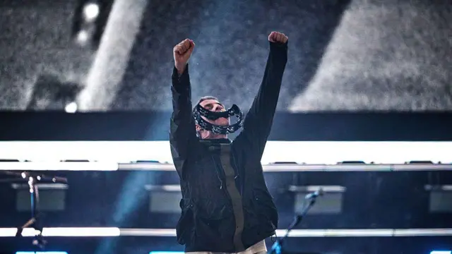 Liam Gallagher segura um pandeiro na boca enquanto levanta os braços no ar