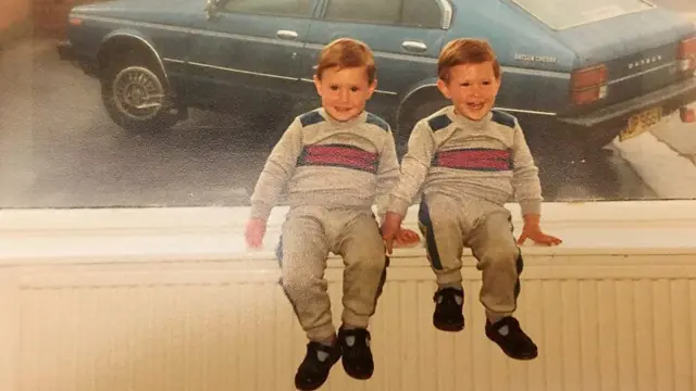 Fotografia antiga mostra dois meninos brancos, gêmeos idênticos, loiros, usando a mesma roupa, sentadosbullsbet appuma plataforma e sorrindo 