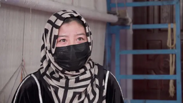 سمیرا با لباس و ماسک سیاه و چادر سیاه و کریمی پشت به یک دستگاه قالین.