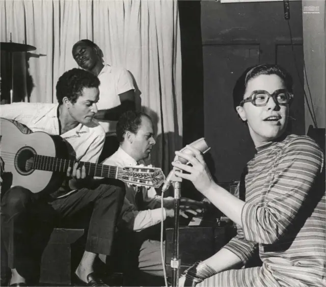 Elis sentada em frente ao microfone, olhando para o lado, com músicos atrás