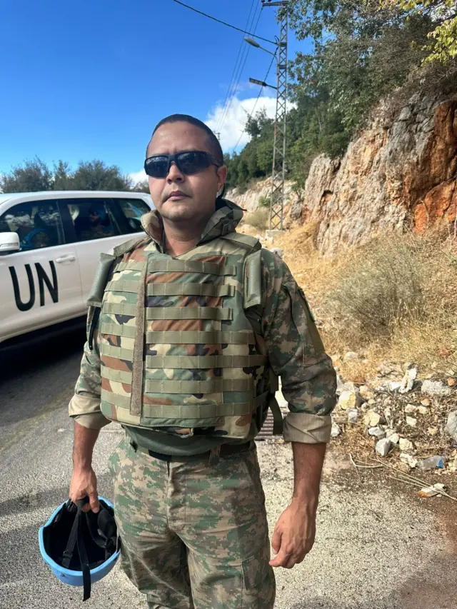 Hamilton de Andrade dos Santos de uniforme, em pé, segurando capacete em uma estrada vazia, ao lado de um carro com os dizeres 'UN'